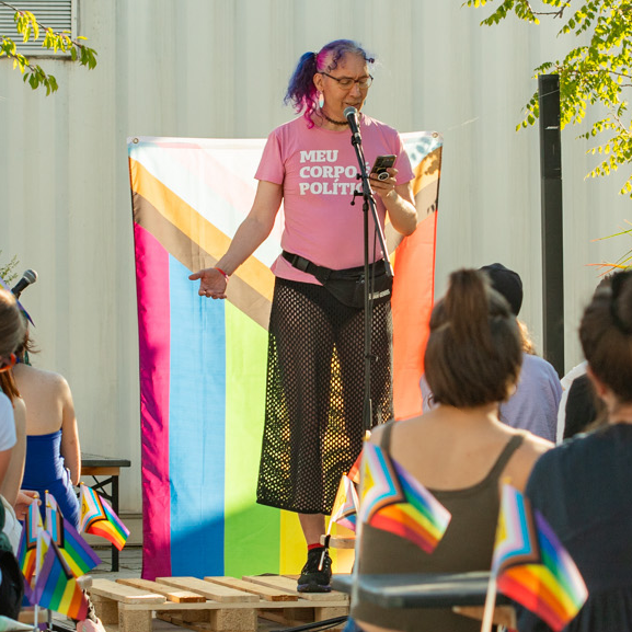 You see Rhonda standing on a stage made of wodden pallets. She's wearing a pink shirt saying "Meo Corpo é Político", a black net-skirt with black panties underneath. Her hair is multi-colored dyed and curly, she's holding a mobile phone and standing in front of a microphone. You see the backs of some people sitting in the audience in front of her. Behind her there is a Progress Pride Flag hanging.