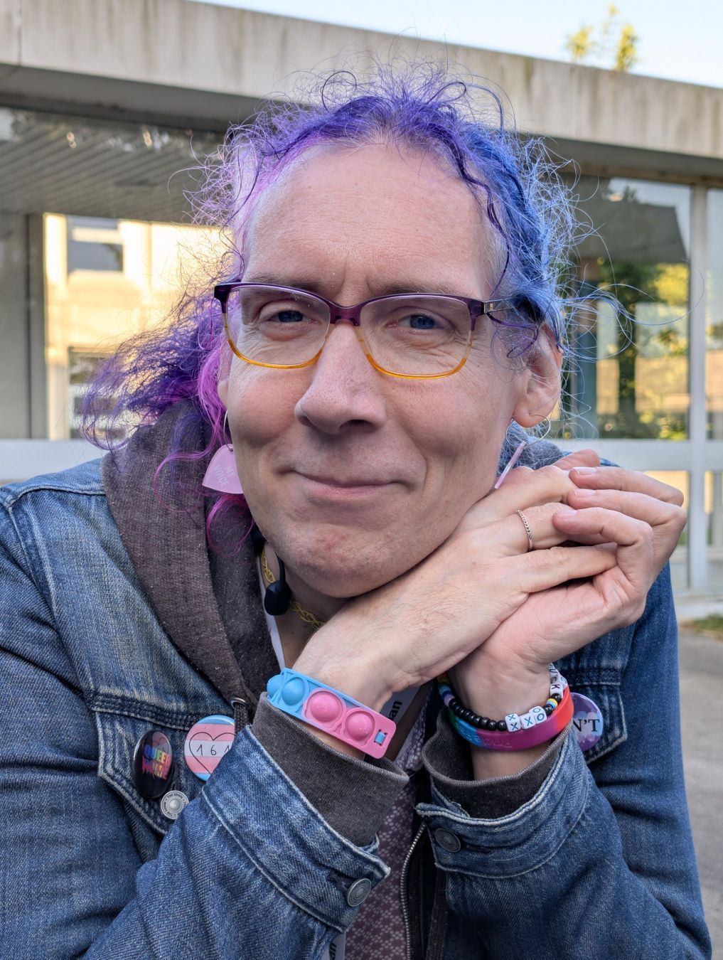 Photo of Rhonda, a white femme with her curly hair in bisexual pride colors. She's wearing a jeans jacket with several buttons on it, one is a trans pride flag with 161 written on it. She's smiling at the camera. Photo of Rhonda, a white femme with her curly hair in bisexual pride colors. She's wearing a jeans jacket with several buttons on it, one is a trans pride flag with 161 written on it. She's smiling at the camera.