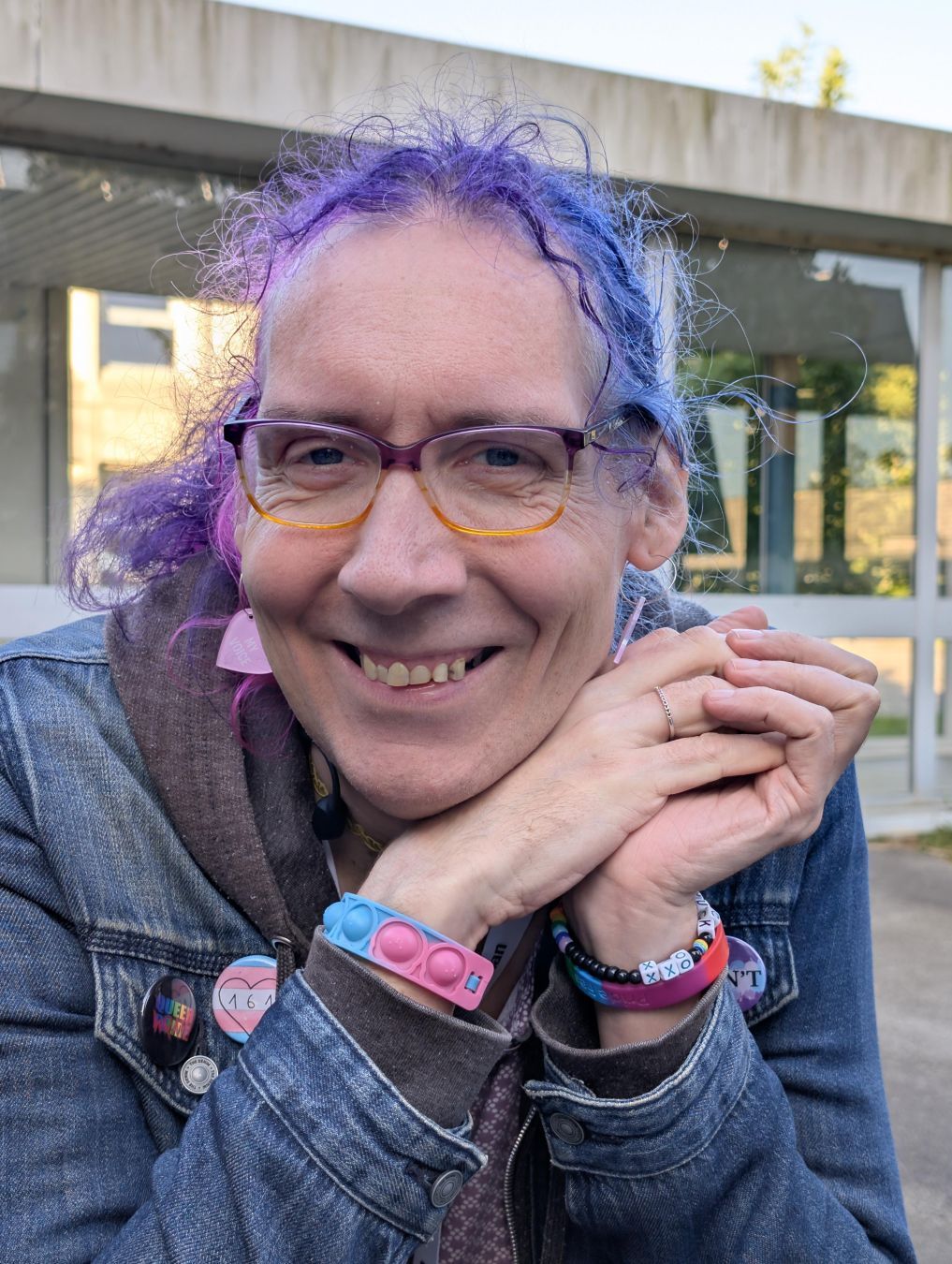 Photo of Rhonda, a white femme with her curly hair in bisexual pride colors. She's wearing a jeans jacket with several buttons on it, one is a trans pride flag with 161 written on it. She's laughing at the camera. Photo of Rhonda, a white femme with her curly hair in bisexual pride colors. She's wearing a jeans jacket with several buttons on it, one is a trans pride flag with 161 written on it. She's laughing at the camera.